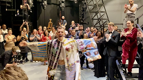 Kyle Brown, wearing the Legacy Robe, runs towards the direction of the camera onstage, while a supportive company cheers him on.