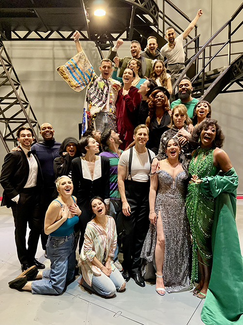 A group of 22 people, many of whom are in costume, and including Brown in the Legacy Robe, stand on a metal staircase on a stage.