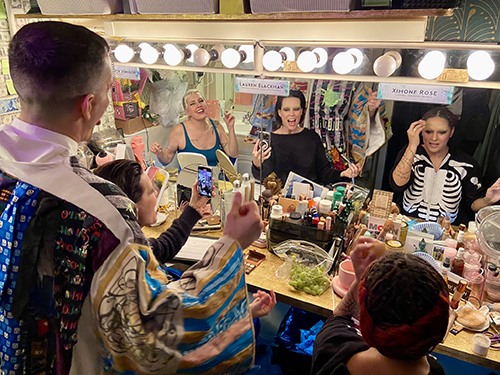 Brown stands with his back to the camera behind three actors, each visible in the mirror of their shared dressing room. The signs above their mirrors indicate that they are Natalie Joy Johnson, Lauren Blackman and Ximone Rose. They are looking excited as they put on their makeup.