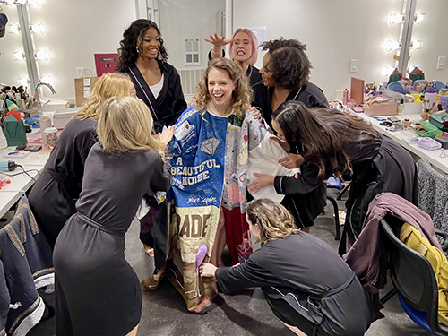 Briggs stands in the Robe in a dressing room. Seven female-presenting people, all wearing dressing robes, lean and gesture towards her, circled around her.