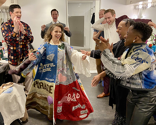 Briggs stands in the Robe in a dressing room. Six male-presenting people circle her and applaud.