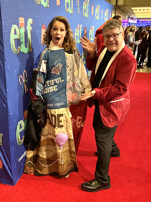 Briggs stands in the Robe on a red carpet in front of a step-and-repeat for the musical 'Elf', looking surprised. Sean Astin, wearing a red jacket, gestures as though to present her.