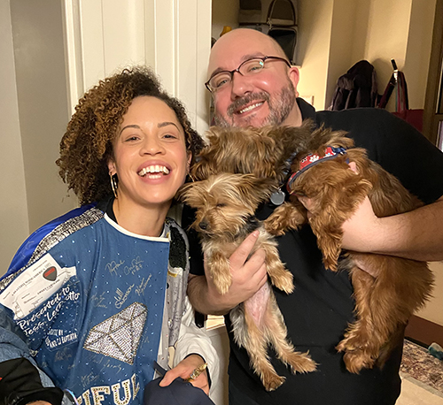 Hutchings stands smiling with a man wearing a black t-shirt. He holds up two small dogs.