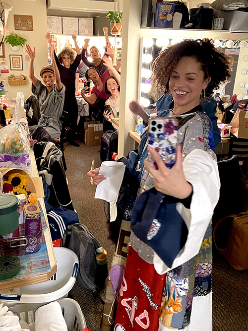 Hutchings poses to take a selfie in a dressing room backstage. Crowding into her frame behind her are five female presenting people.