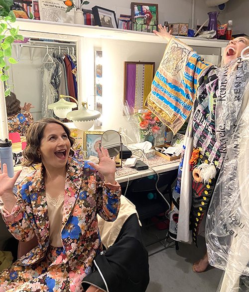 Shaina Taub is seated at her dressing room table, throwing her hands up excitedly. Scott stands above her in the Robe, throwing her arms up in the air.