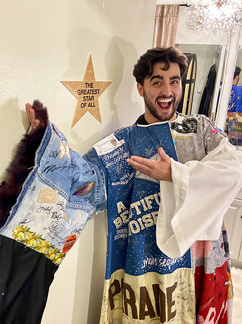 Giuseppe Bausilio stands wearing the Robe, grinning and gesturing to a star on a dressing room door that reads 'The Greatest Star of All'.